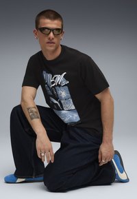 Mand iført mørke solbriller, sort grafisk T-shirt, mørke jeans og blå sneakers knælende med tatovering på venstre underarm mod en grå baggrund.