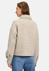 Veste beige texturée avec un large col, des épaules tombantes et des poignets boutonnés, associée à un jean en denim bleu.