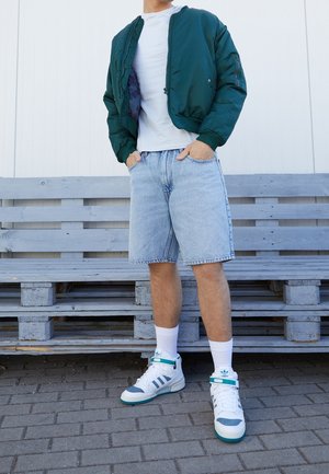 Veste aviateur verte avec une fermeture éclair noire, t-shirt blanc, short en denim bleu clair, et baskets blanches avec des accents turquoise et des détails gris.