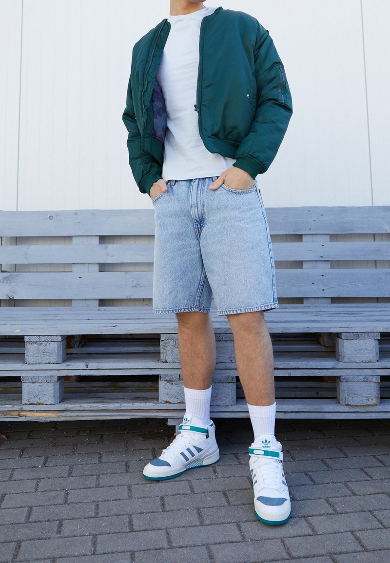Veste aviateur verte avec une fermeture éclair noire, t-shirt blanc, short en denim bleu clair, et baskets blanches avec des accents turquoise et des détails gris.