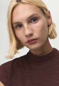 Close-up of a person with short blonde hair, wearing a maroon high-neck top. Features freckled skin and blue eyes. Smooth texture, simple design.