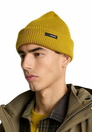 Yellow ribbed beanie hat with a pointed top and black label. Model wears a green jacket over a plaid shirt and yellow shirt underneath.