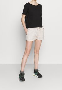 Černý top s krátkým rukávem a kulatým výstřihem, kombinovaný s béžovými šortkami na zavazování. Model má na nohou černé sportovní boty se zelenými akcenty.