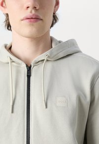 Felpa con zip grigia chiara realizzata in tessuto testurizzato, con zip frontale e sottile patch del logo "BOSS" sul petto.