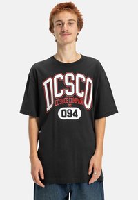 Čierne bavlnené tričko s veľkým červeným a bielym grafikou s nápisom "DC Shoe Company" a "094", vo voľnom a uvoľnenom strihu s krátkymi rukávmi.