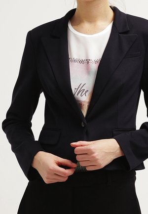 Kvinde, der knappes en tætsiddende sort blazer over en hvid T-shirt med grafik, vist fra hals til hofter mod en ensfarvet baggrund.