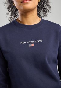 Donkerblauwe sweatshirt van katoen, met de tekst "NEW YORK STATE" in witte letters en een kleine afbeelding van de Amerikaanse vlag. Zachte textuur.