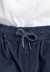 Pantaloni corti in corduroy blu scuro con cintura elastica, dotati di un cordoncino e terminali metallici. Camicia bianca parzialmente visibile sopra.