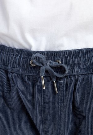 Shorts en velours côtelé bleu marine avec une taille élastique, dotés d'un cordon de serrage et d'embouts en métal. Chemise blanche partiellement visible au-dessus.
