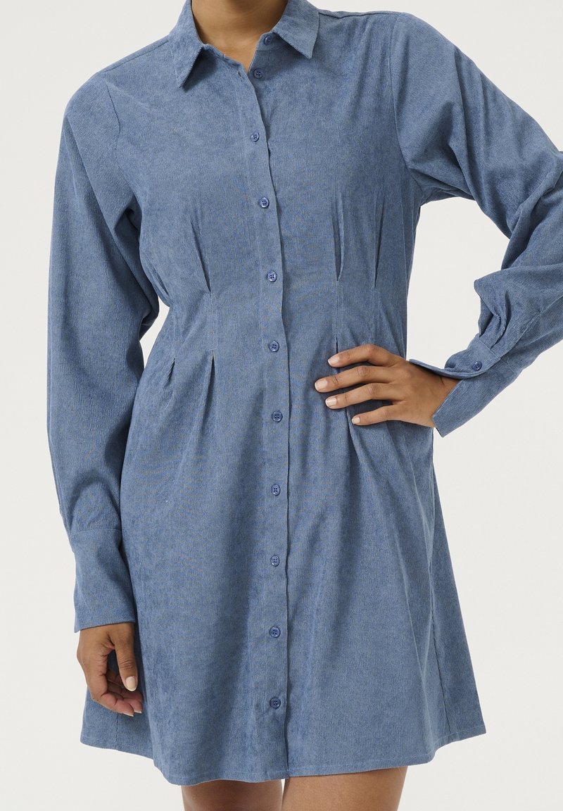 Femme portant une robe en velours côtelé bleue à manches longues, boutonnée, avec col et taille cintrée, debout, une main sur la hanche.
