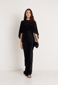 Černé maxi šaty s pláštěm na překrytí. Hladká látka, přiléhavý střih a minimalistický design. Doplněno malou černou kabelkou.