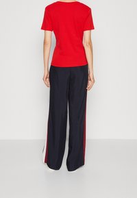 Person klädd i en röd kortärmad skjorta och lösa svart byxor med röda och vita sidränder, stående mot en enhetlig ljus bakgrund.