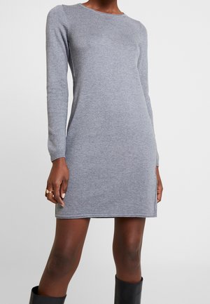 Femme portant une robe en maille grise ajustée, à manches longues et longueur genou, associée à des bottes noires, debout devant un fond clair uni.