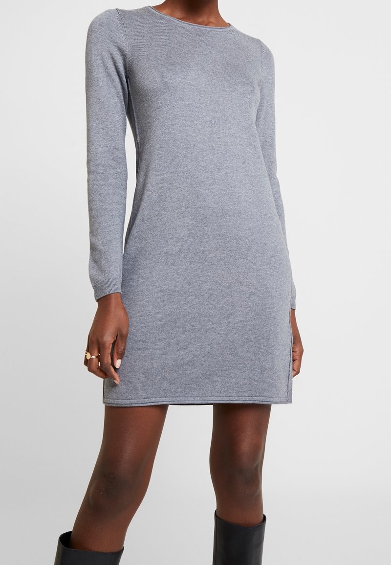 Femme portant une robe en maille grise ajustée, à manches longues et longueur genou, associée à des bottes noires, debout devant un fond clair uni.