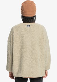 Beige fleecepullover med lös passform, texturerad yta och vida ärmar. Har en logotyp på baksidan och rund halsringning. Bärs med en brun stickad mössa.