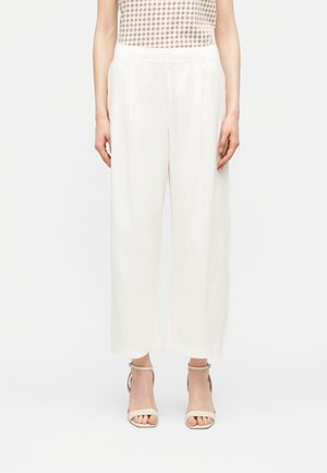 Γυναίκα που φοράει λευκό παντελόνι με φαρδιά μπατζάκια, μπεζ αμάνικο τοπ με καρό σχέδιο και μπεζ σανδάλια με λουράκι στον αστράγαλο, στέκεται μπροστά σε λευκό φόντο.