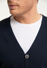 Mörkblå v-ringad cardigan i stickad kvalitet, med en liten silverknappdetalj vid halsen över en vit t-shirt.