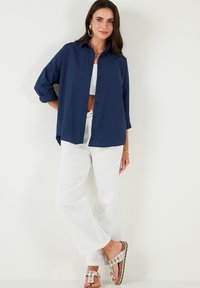 Camicia navy blu con bottoni, a maniche lunghe e taglio rilassato. Abbinata a pantaloni bianchi a gamba larga e sandali con accenti argentati. Tessuto liscio.