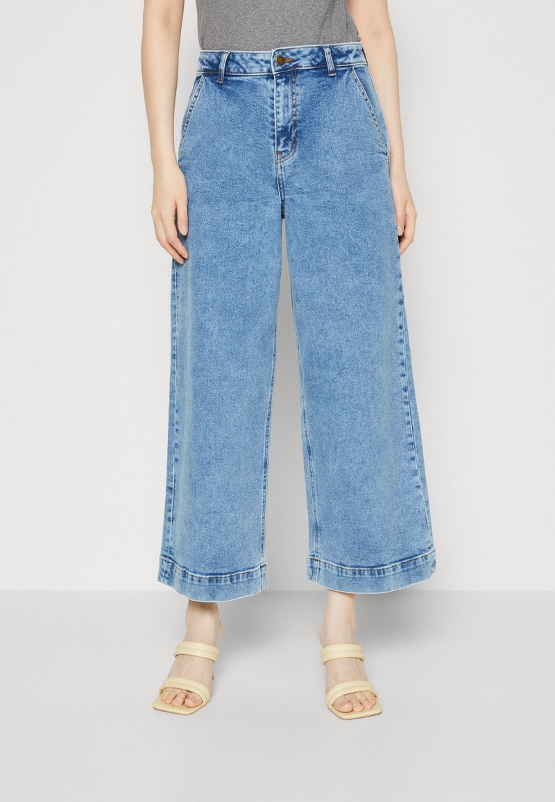 Nainen, jolla on yllään vaaleansiniset korkeavyötäröiset leveälahkeiset farkut ja beigeet avokkaat, joissa on kaksoishihnat, seisoo yksinkertaista taustaa vasten.