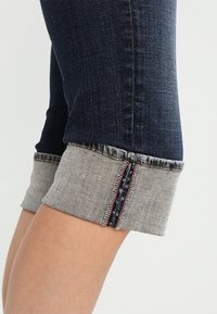 Tumman siniset farkut harmailla käänteillä, joissa on kontrastiväriset punaiset ja tummansiniset saumat, korostaen tekstuuria ja muotoilua.