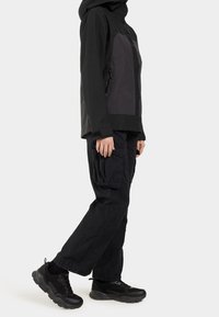 Veste imperméable noire avec capuche, dotée d'un panneau avant gris et de poches zippées, associée à un pantalon cargo noir ample et des baskets noires.