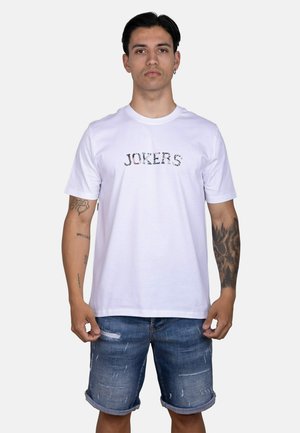 Hombre con camiseta blanca con texto "JOKERS" y pantalones cortos de mezclilla rasgados, de pie contra un fondo claro y liso.