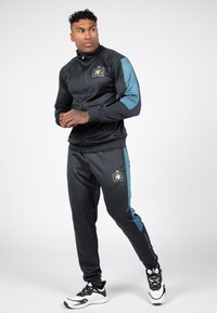 Zwarte ritsjack met aquamarijnaccenten op de mouwen, gecombineerd met bijpassende zwarte joggers met zijstrepen en logo. Het materiaal lijkt glad en sportief.