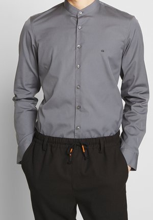 Mann trägt ein eng anliegendes graues Hemd mit Stehkragen und Knopfleiste, versehen mit dem "ck"-Logo, und schwarze Kordelhose mit orangefarbenen Akzenten, Hände in den Taschen.