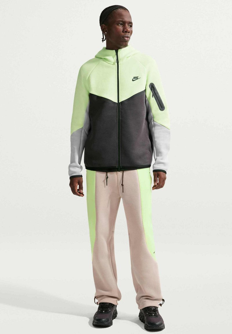 Mand iført neon grøn, sort og grå Nike-hættetrøje med beige og neon grønne bukser, stående og kiggende mod højre på en hvid baggrund.