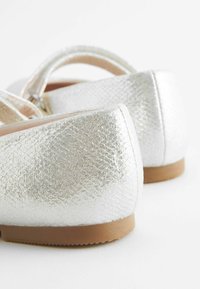 Silverfärgade metalliska ballerinas med en texturerad yta, rundad bakdel och beige gummisålar. Inkluderar ett tunt band för säker användning.