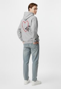 Felpa grigia con un design a cuore rosso sulla schiena, con delle forbici nere. Abbinata a jeans azzurri chiari e sneakers bianche.