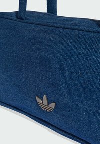 Denim tote bag in dark blue with a textured surface. Features two handles and a metallic logo accent on the front.