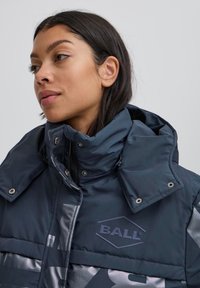 Chaqueta acolchada azul marino con cuello alto, con un logo plateado brillante y detalles. La textura parece suave y aislante.