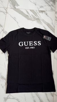 Juodas medvilnės marškinėliai trumpomis rankovėmis, ant kurių priekyje yra baltas "GUESS" logotipas ir tekstas "EST.1981". Prezentacija iš viršaus.