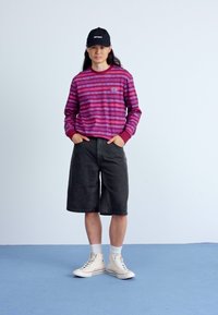 Camisa de rayas morada y rosa de manga larga, pantalones cortos negros, zapatillas blancas y gorra negra. El modelo está de pie con las manos en los bolsillos sobre un suelo azul.
