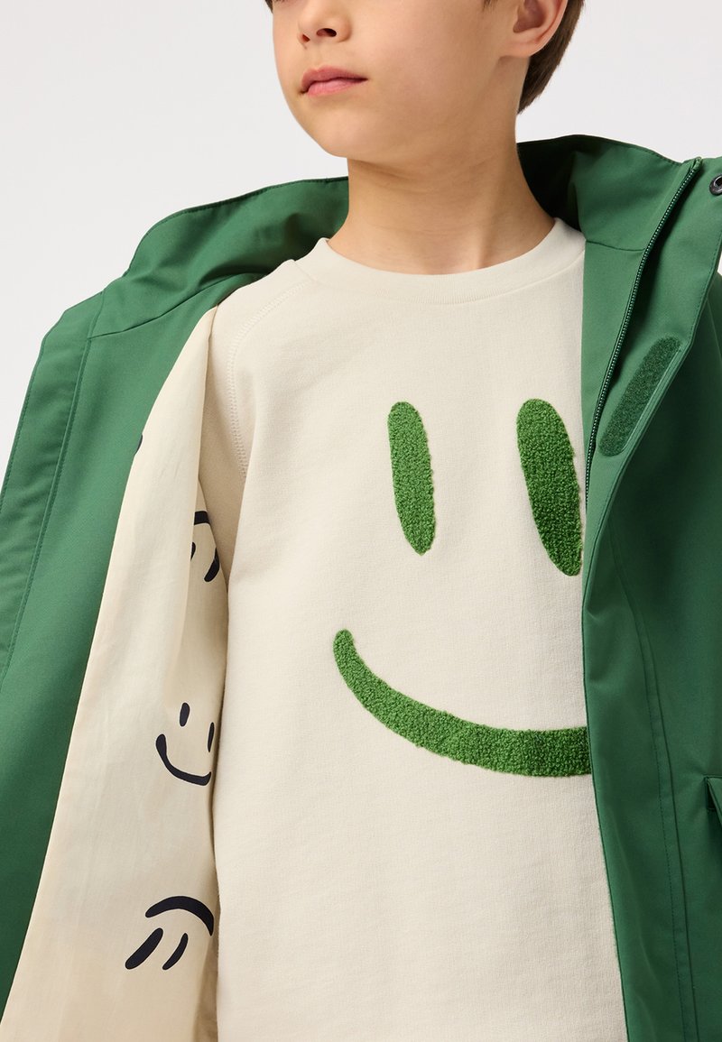 Felpa in cotone beige con design di faccina sorridente verde in morbida pelliccia, abbinata a un giubbotto impermeabile verde con texture liscia e tasche funzionali.