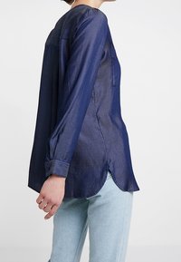 Camicia a maniche lunghe blu navy in un tessuto leggero e liscio. Presenta un orlo arrotondato e una tasca sulla schiena. Polsini con bottoni.