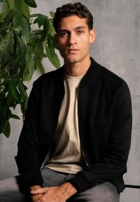 Veste bomber noire en suédine avec un col côtelé, assortie à un t-shirt beige clair. Jeans gris visibles. Plante en arrière-plan.