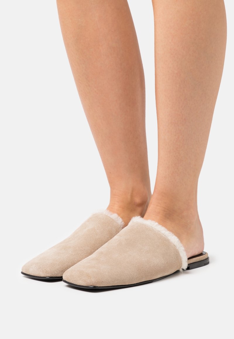 Chaussons beiges en daim, sans talon, doublés de fausse fourrure blanche, portés pieds nus sur un fond blanc.