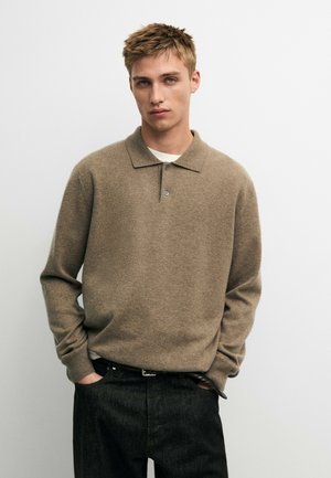 Junger Mann mit kurzen blonden Haaren, der einen braunen langärmligen Polo-Pullover und dunkle Hosen trägt, mit den Händen in den Taschen vor weißem Hintergrund.