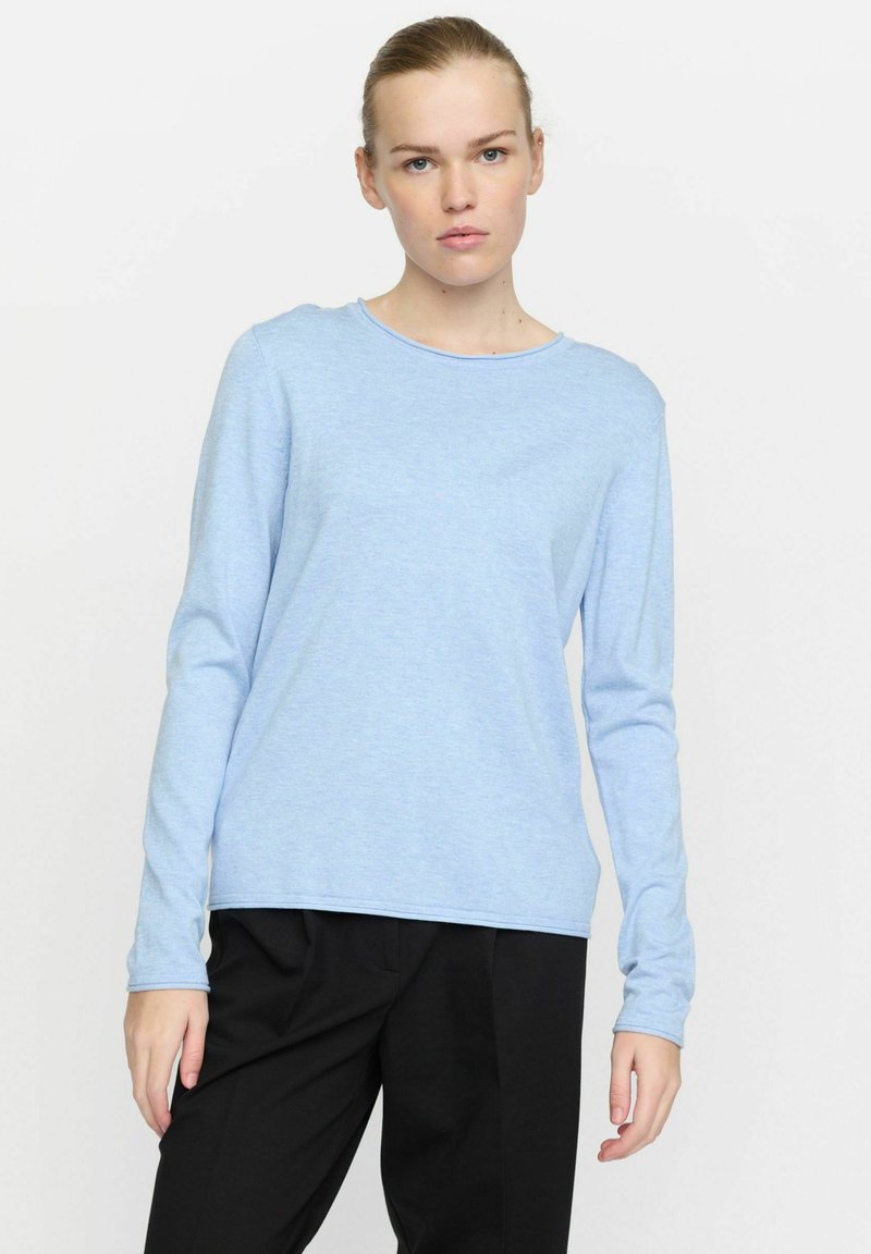 Camicia azzurra chiara a maniche lunghe con scollatura rotonda, texture morbida e vestibilità rilassata, abbinata a pantaloni neri. Nessun motivo o accento particolare.