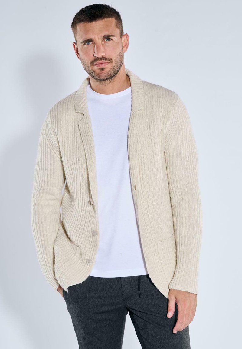 Beige ribbed cardigan with shawl collar, button front, and two pockets, layered over a white t-shirt, paired with dark gray pants.