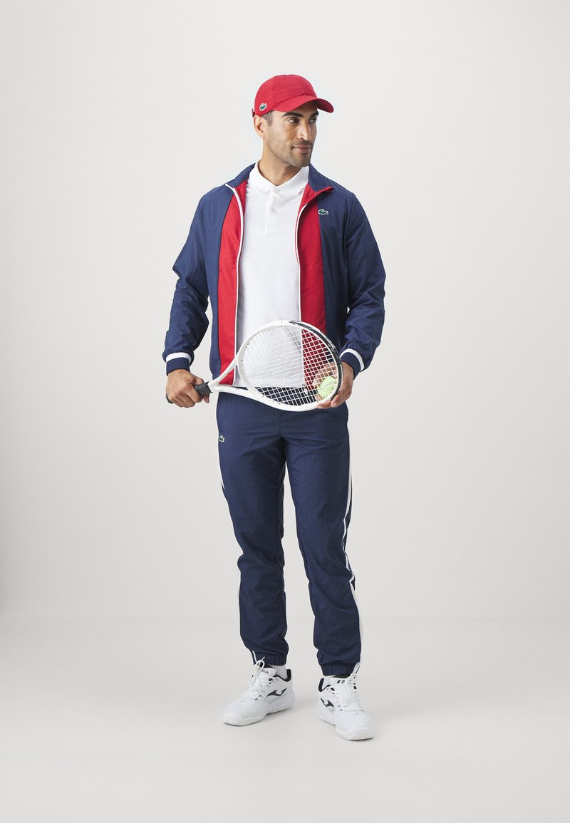 Homme portant une casquette rouge et un survêtement bleu marine et rouge, tenant une raquette de tennis et une balle, se tenant devant un fond clair uni.