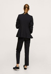 Femme avec des cheveux attachés portant un blazer noir et un pantalon, se tenant de dos, assortie de baskets noires et beiges sur un fond uni.
