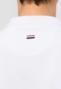 Dos d’un polo blanc porté par une personne, montrant un petit logo rayé horizontal rouge et marine sous le col.
