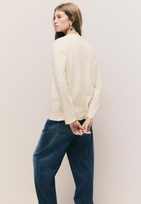 Camisă lungă cu mâneci lungi, de culoare crem, cu o croială lejeră, purtată cu pantaloni din denim negru, lejeri. Design simplu, fără modele sau accesorii vizibile.