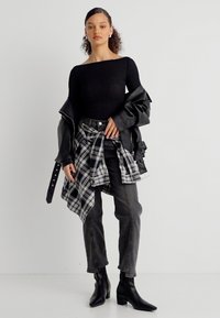 Γυναίκα όρθια με μαύρο μπλουζάκι χωρίς ώμους, μαύρο δερμάτινο μπουφάν, μαύρο καρό πουκάμισο δεμένο στη μέση, σκούρα τζιν και μαύρες μποτάκια αστραγάλου.