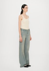 Top in tessuto a coste beige con spalline larghe, abbinato a jeans a gamba larga di colore blu chiaro. Il modello indossa scarpe slip-on nere e presenta orecchini a cerchio piccoli.