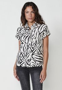 Camisa de botones de manga corta en beige con estampado de cebra negro. Presenta dos bolsillos frontales y un cuello tipo clásico. Tejido suave y ligero.