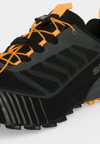 Chaussure de trail noire et grise avec des lacets orange vif et une languette, présentant une semelle robuste et une tige Gore-Tex imperméable.